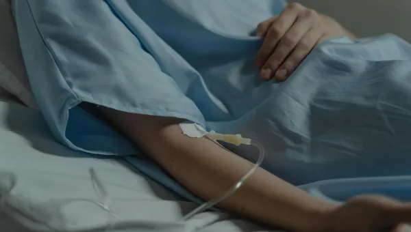 A close-up of a hospital patient's arm with an IV line and a light blue gown covering the body in a bed.
