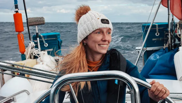A person stands at the wheel of a small sailing boat on open water. The person wears a knitted hat with a pom-pom and a padded jacket.