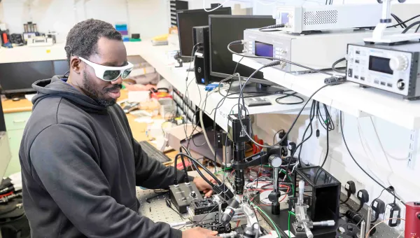 Student adjusts photonics laboratory equipment while wearing protective laser safety glasses.