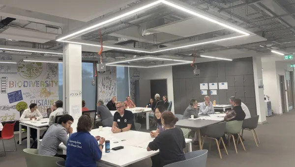 People collaborate around tables in a modern, brightly lit meeting room with sustainability-themed wall artwork.