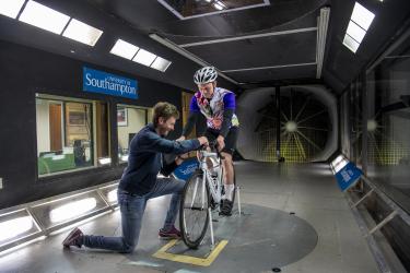 Man on bike in wind tunnel with man adjusting his wheel.