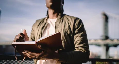 A Black man writes on a notepad as he stands outside, a blue sky and a bridge behind 