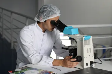 A researcher in a white lab coat looks through a microscope.