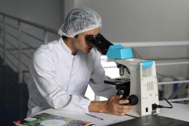 A researcher in a white lab coat looks through a microscope.