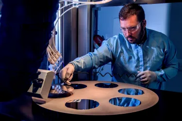 A bearded young scientist in protective overalls and glasses works with a flame hydrolysis deposition (FHD) system