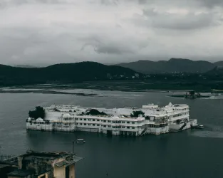 The Taj Lake Palace in Udaipur, Rajasthan, India. It is a rainy day and there are dark clouds surrounding the hills in the distance.