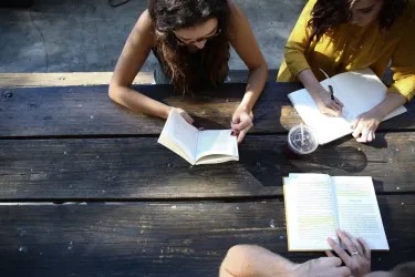 Three people gathered around at a picnic table while reading books and writing notes