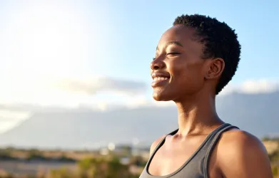 Person with short hair smiling with eyes closed against a bright sky background.