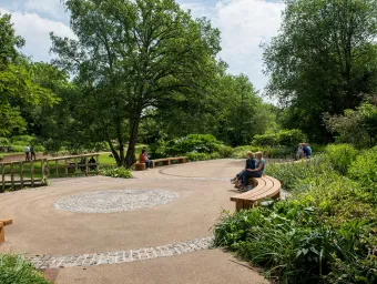 People relaxing on benches surrounded by trees, plants and parkland on a sunny day.