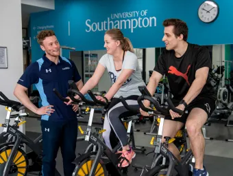Two students working out on exercise bikes in a gym, assisted by a personal trainer.