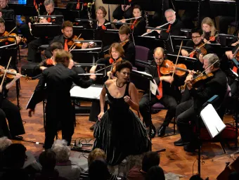 Female opera singer performing, surrounded by members of an orchestra.