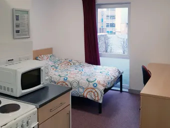 View of a studio flat showing a double bed and wooden desk. A kitchen area with microwave and hob is also partially visible.