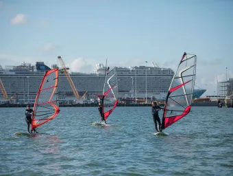 Three people windsurfing beneath blue skies on a sunny day. A large cruise ship is visible behind them, docked in port.