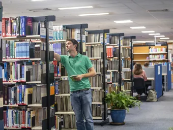 A student selects a book from a shelf while other students work at desks in the background