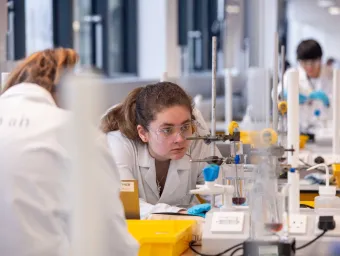 A student in a lab coat crouches down to closely monitor a chemistry experiment being conducted in a large piece of glassware.
