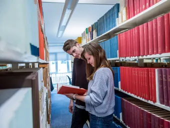 Two students in a narrow library aisle, surrounded by shelves of books on both sides. They are reading a book pulled from one of the shelves.