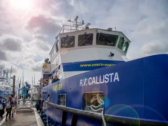 An industrial looking boat emblazoned with the name RV Callista, moored beside a wooden dock, beneath a sunny sky.