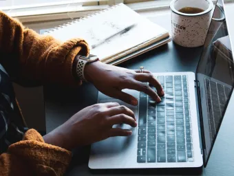 Fingers typing on a laptop with notepad, pen and mug in the background