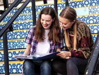 Two students sitting together on the stairs outside a University building, comparing their notes from a recent lecture.