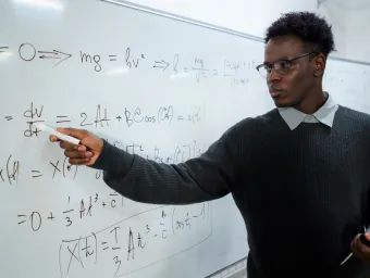 A maths teacher writes on a white board and looks intently at the equations. We imagine he is in front of a classroom of pupils.