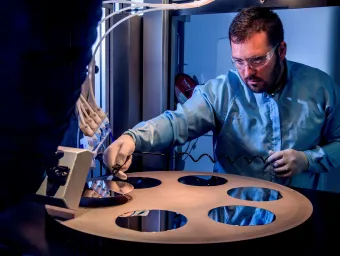 A bearded young scientist in protective overalls and glasses works with a flame hydrolysis deposition (FHD) system
