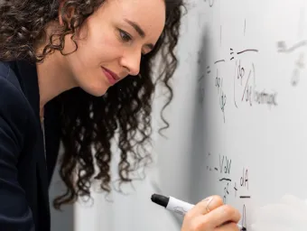 Close-up of a young woman with long, curly dark hair writing equations on a whiteboard