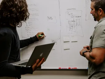 Two people work together on a white board, looking at what one is writing on the board, which is already full with text