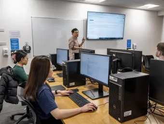 Three students in front of monitors in a classroom listen as a lecturer makes a point