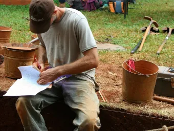 An archaeologist sits on the edge of a freshly dug trench, making notes on a clipboard
