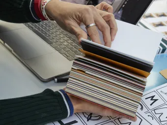 Two hands flicking through a swatch book. Hand drawn sketches in the foreground, with contact sheets and a laptop