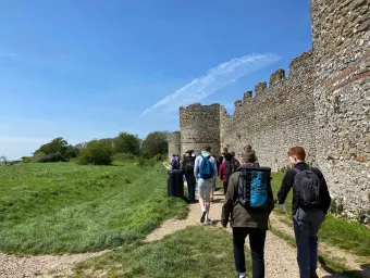 A group of history students walk beside the outside wall of the Hampshire medieval fortress Portchester Castle, as part of a field trip. The 2 students closest to us are in discussion.