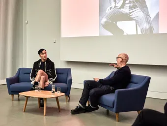 Charles Jeffrey and Professor Jonathan Faiers sit on blue sofas with a small table between them. Jonathan is facing away from the camera and has an arm raised as he speaks. Charles is facing Jonathan and appears to be concentrating on what Jonathan is saying. There is a projector behind them showing a person wearing a pinstripe suit.