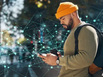 A man in a yellow beanie and olive-green sweater, engrossed in a smartphone, holding a disposable coffee cup, and wearing a backpack. He is standing against a backdrop of an abstract digital network design with interconnected nodes and lines.