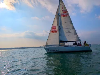 A yacht with 2 sails and a training crew sailing out on the water. The  sky has clouds and an orange eveningglowsky