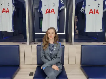Emma Green wearing a Tottenham Hotspur tracksuit, is sitting on a chair with 3 Tottenham Hotspur shirts hanging behind her.