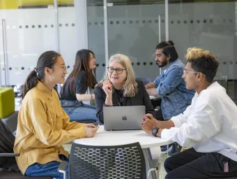 Three people sat a desk talking. The person in middle has a laptop open on the desk.