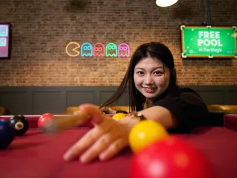 A student playing pool in a students' union bar. The student has long dark hair, and the background shows a brick wall with televisions and a neon sign in the shape of characters from the game Pac-Man.