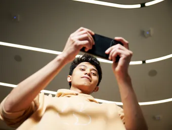 A student is looking down at the camera while using their smartphone, which they are holding horizontally. The student is wearing a pale yellow hoodie, and stood in a corridor which is well-lit by several fluorescent lighting strips.