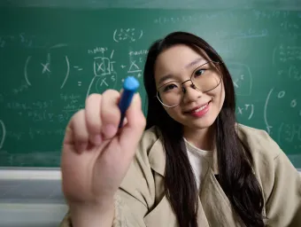 A student using a white marker pen to write a mathematical equation on a glass surface. They are wearing glass and a beige jacket, and they are stood in front of a blackboard which also features a mathematical equation.