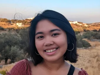 Amatta Mirandari smiling at the camera. She is wearing a dark red muslin top. There is scrubland and blue sky in the background.