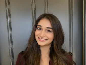 Disha Mehta smiling at the camera, her dark brown hair is long and wavy. It falls over her burgundy jacket.