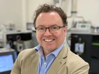 Professor Max Crispin smiling at the camera. He is standing in a lab with a lot of technical equipment in the background.