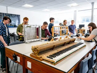 A group of students sits around a long table in a classroom, where a piano keyboard has been placed for an experiment.