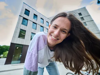 A smiling student stood outside the Centenary Building, also known as Building 100, on the University of Southampton's Highfield campus. They have long hair and wearing a purple and white shirt with blue jeans.