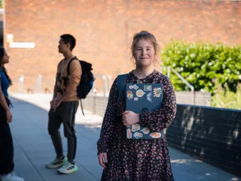 A student smiling while stood outside and holding a closed laptop. There are two more students talking to each other in the background.