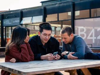 Three students sat on a bench outside the University of Southampton's students' union. They are a looking at a smartphone held by the student in the middle.