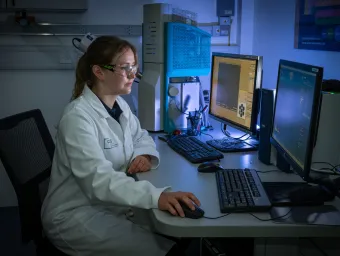 A Chemistry and Chemical Engineering technician in a lab, wearing a lab coat and goggles while sat at a desk using a computer.
