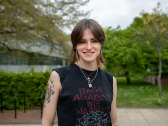 A student stands smiling outside the Physics building dressed casually in a sleeveless band T-shirt.