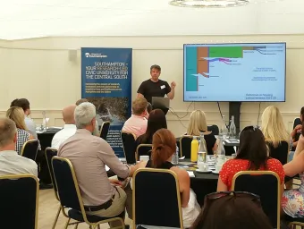 A man standing next to a presentation screen showing an illustration of outcomes in housing judicial reviews. There is a seated audience in front of him. 