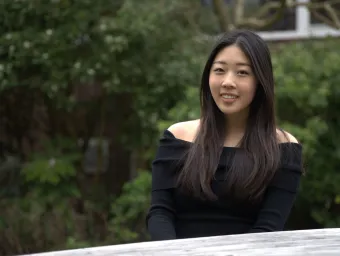 Student sitting outdoors with green shrubs behind her. She is smiling at the camera.